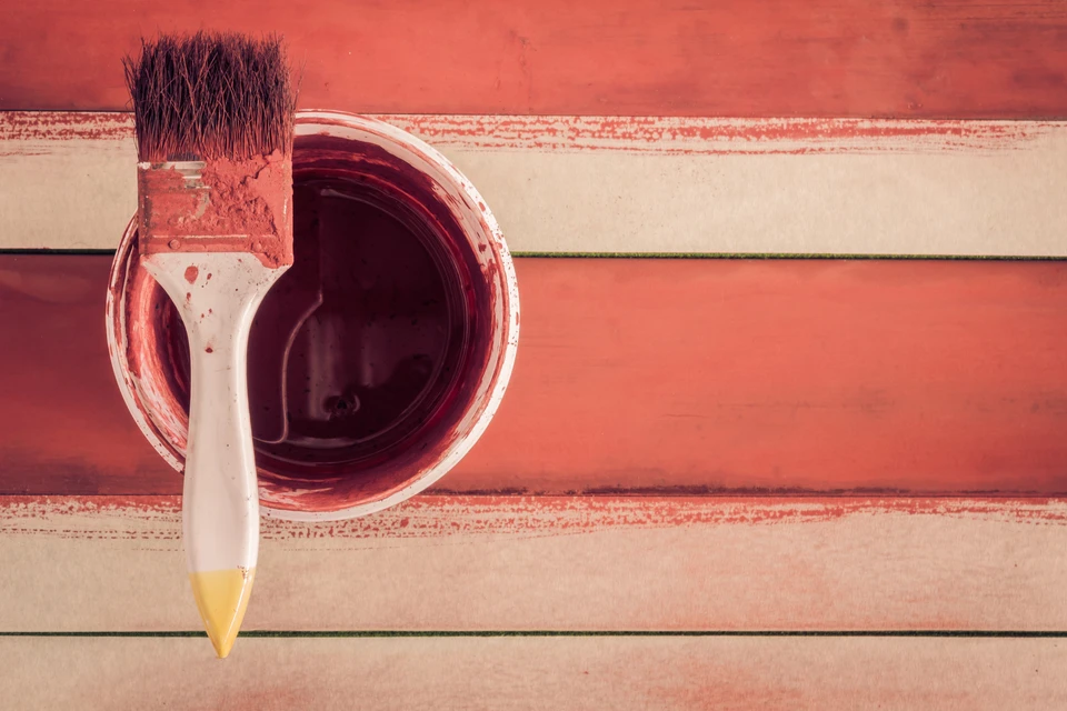 Scène d'intérieur montrant un pinceau plat à manche blanc et virole dorée trempé dans un pot de peinture rouge bordeaux, posé sur des planches de bois peintes en rouge et blanc avec finitions patinées