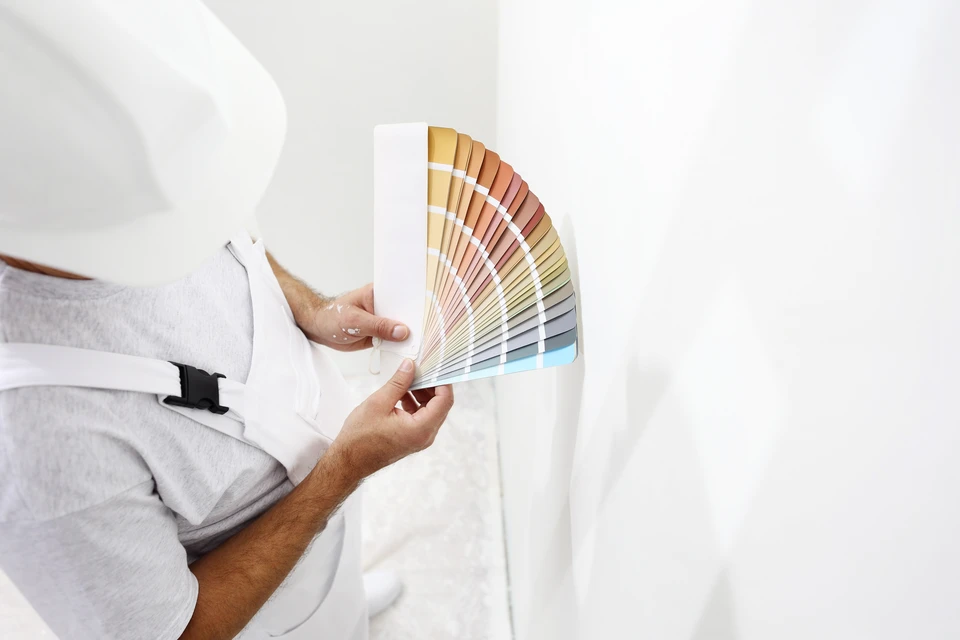 Personne tenant un nuancier de couleurs en éventail contre un mur blanc, évaluant des teintes pour travaux de peinture intérieure