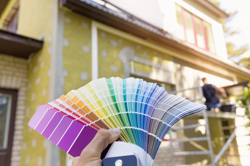 Une main tenant un éventail de nuanciers de couleurs devant une façade de bâtiment jaune en rénovation, avec échafaudages visibles