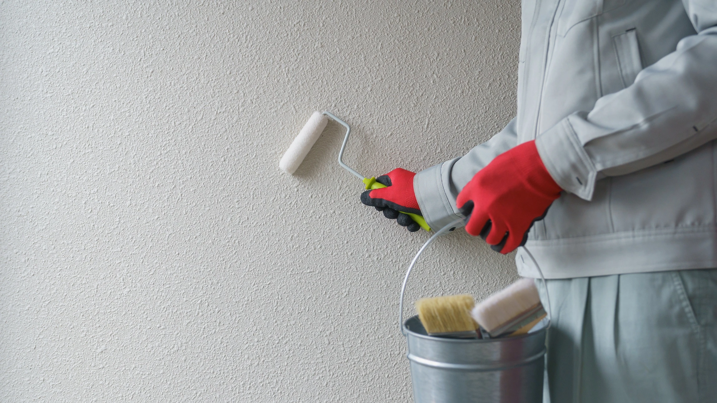 Peintre professionnel en combinaison grise et gants rouges appliquant de la peinture murale avec un rouleau sur mur texturé beige